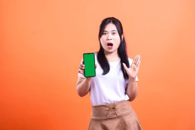 Şaşırmış bir kadın elinde yeşil ekranlı akıllı bir telefon aleti tutuyor. Onun ağzı açık ve bir eli havada şok olmuş bir ifadesi var. Turuncu arka planda izole edilmiş..