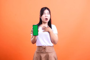 Beyaz tişört ve kahverengi etek giyen bir kadın, turuncu arka planda izole edilmiş, şaşırmış bir ifadeye sahip yeşil ekranlı bir telefon tutuyor..