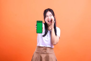 Beyaz gömlek ve kahverengi etek giyen, elinde yeşil ekranlı siyah bir cep telefonu olan Endonezya 'lı genç bir kadın, turuncu bir arka planda izole edilmiş mutlu bir ifadeyle yüzünü çeviriyor..