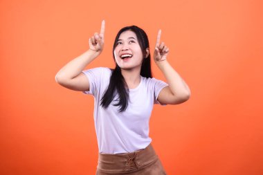 Siyah saçlı genç bir kadın, turuncu arka planda izole bir şekilde yukarı bakarken işaret parmağıyla işaret ediyor. Beyaz bir tişört ve kahverengi bir etek giyiyor..