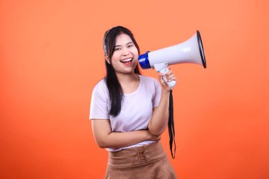 Siyah saçlı, beyaz tişörtlü ve kahverengi etekli bir kadın elinde mavi kenarlı beyaz bir megafon tutuyor. Gülümsüyor ve parlak turuncu bir geçmişle mutlu görünüyor..