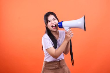 Bir kadın ağzında beyaz bir megafon tutuyor ve konuşuyor gibi görünüyor. O, beyaz bir gömlek ve kahverengi bir etek giyiyor ve turuncu bir arka planda izole edilmiştir..