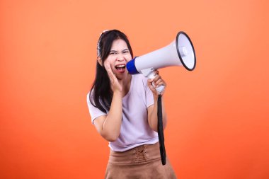 Bir kadın parlak turuncu arka planda megafona bağırıyor. O, beyaz bir gömlek ve kahverengi bir etek giyiyor ve megafonu iki eliyle tutuyor. Önüne bakıyor ve ağzı açık..