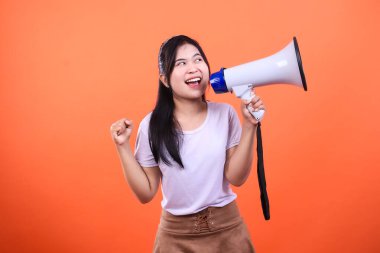 Bir kadın elinde mavi başlıklı beyaz bir megafon ve turuncu arka planda izole edilmiş siyah bir tutam tutuyor. Gözlerini kaçırıyor ve yumruğunu kaldırıyor..