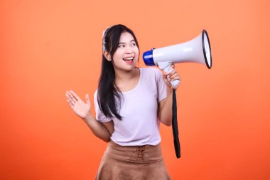 Solda görülen genç bir kadın siyah kayışlı beyaz bir megafon tutuyor ve tek eliyle sallıyor. Açık mor bir gömlek ve kahverengi bir etek giyiyor. Görüntü turuncu bir arkaplanda izole edilmiş..