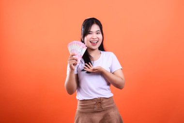 Bir kadın fatura hayranı ve gülümsüyor. O, beyaz bir tişört ve kahverengi bir etek giyiyor. Turuncu bir arka planda izole edilmiş..