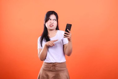 Hoşnutsuz bir ifade takınan bir kadın turuncu arka plan önündeki siyah akıllı telefonunu işaret ediyor. Beyaz bir tişört ve kahverengi bir etek giyiyor..