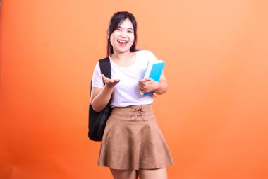 Genç bir öğrenci turuncu bir arka planda, beyaz bir gömlek, kahverengi bir etek ve siyah bir sırt çantası giyerek ve elinde mavi bir kitap tutarak tek başına duruyor. Gülümsüyor ve mutlu görünüyor..