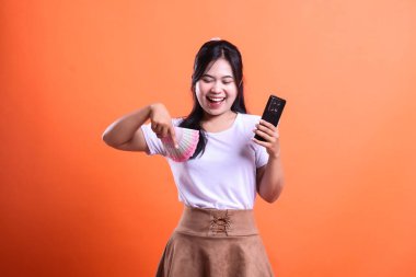 Gülümseyen bir kadın bir elinde Endonezya Rupiah banknotları, diğer elinde turuncu arka planda izole edilmiş akıllı bir telefon cihazı tutar..