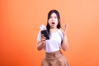 Şaşırmış bir kadın elinde para dolu siyah bir cüzdan tutuyor. Beyaz gömlek ve kahverengi etek giyiyor ve ağzı açık, turuncu arka planda izole bir şekilde şokta..