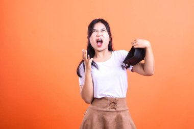 Bir kadın elinde boş bir siyah çanta tutuyor ve öfkeli bir tepki gösteriyor. Beyaz bir tişört ve kahverengi bir etek giyiyor, turuncu arka planda izole edilmiş..