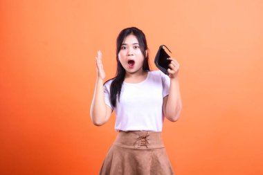 Bir kadın elinde boş bir cüzdan ve şok olmuş bir ifadeyle turuncu bir arkaplanda duruyor. Diğer elini kaldırdı ve sürpriz ve finansal sıkıntıyı önerdi.