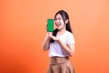 Genç bir kadın elinde yeşil ekranlı akıllı bir telefon tutuyor, diğer eliyle ekrana doğru işaret ediyor. Turuncu arka planda izole edilmiş beyaz ve kahverengi bir etek giyiyor..