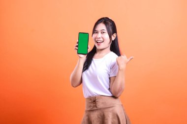 Gülümseyen bir kadın yeşil ekranlı bir cep telefonu tutuyor ve başparmağıyla işaret ediyor. Beyaz bir gömlek ve turuncu bir arka planda izole edilmiş kahverengi bir etek giyiyor..