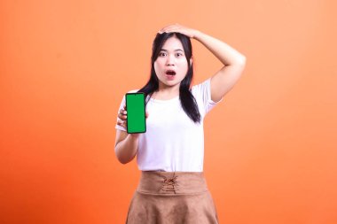 Şaşırmış bir suratı olan bir kadın elinde yeşil ekranlı bir akıllı telefon tutuyor. Bir eli kafasında ve diğer elinde turuncu arka planda izole edilmiş bir telefon var..