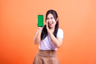 Asyalı bir kadın yeşil ekranlı akıllı bir telefonu tutarken gülümsüyor. Elleri ağzının yanında beyaz tişört ve kahverengi etek giyen bir şey anons ediyor. Turuncu bir arka planda izole edilmiş.