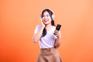 Uzun siyah saçlı, beyaz tişörtlü, kahverengi etekli ve beyaz kulaklıklı bir kadın, siyah bir telefon tutuyor ve turuncu bir arka plana gülümsüyor..