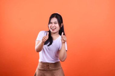 Siyah saçlı, beyaz tişörtlü ve kahverengi etekli bir kadın gülümsüyor ve turuncu bir arka planın önünde iki parmağını kaldırıyor..