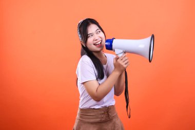 Genç bir kadın konuşurken ya da bağırırken iki eliyle bir megafon tutuyor. Siyah çizgilerle beyaz ve mavi, arka plan turuncu.