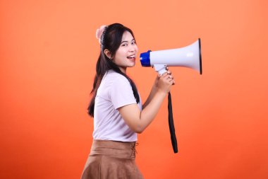 Genç bir kadın elinde megafonla konuşurken beyaz bir megafon tutuyor, arka plan turuncu bir renk ve turuncu bir arka planda izole edilmiş..