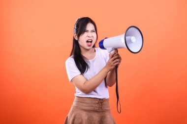 Genç bir kadın, turuncu arka planda izole edilmiş siyah bir kordonla beyaz bir megafona bağırıyor. O beyaz bir tişört ve kahverengi bir etek giyiyor..