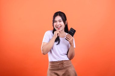 Bir kişi akıllı bir telefon tutuyor ve gülümsüyor, turuncu bir arka planda izole halde. Kişi beyaz bir gömlek ve kahverengi bir etek giyiyor. Arka plan turuncu..