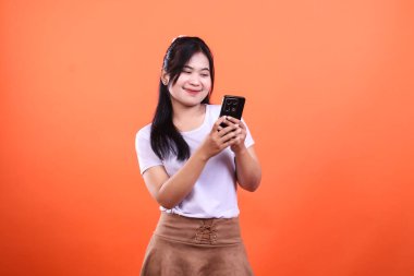 Uzun siyah saçlı bir kadın elindeki cep telefonuna bakıyor. O, beyaz bir tişört ve kahverengi bir etek giyiyor. Bu resim turuncu bir arkaplanda izole edilmiştir..