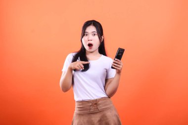 Şaşırmış bir kadın elinde telefon tutarken, turuncu bir arka planda kendini işaret ediyor..