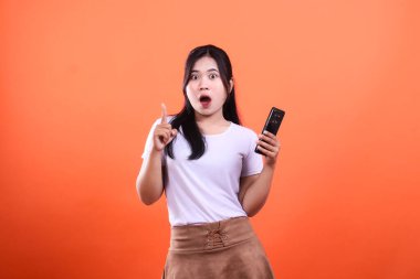 Şaşırmış bir kadın elinde turuncu arka planda bir telefon tutuyor.