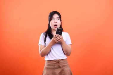 Yanlara bakarken cep telefonu tutan genç bir kadın, ağzı açık, sanki şaşırmış ya da şok olmuş gibi, turuncu bir arka planda izole edilmiş..