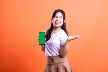 Beyaz gömlekli ve kahverengi etekli bir kadın yeşil ekranlı bir cep telefonu tutuyor ve diğer eliyle hareket ederken gülümsüyor. turuncu arkaplanda izole.