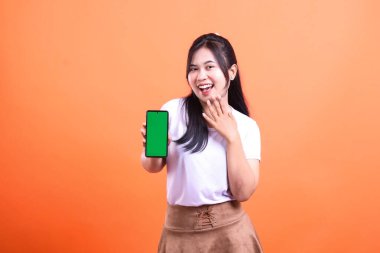Gülümseyen bir kadın yeşil ekranlı bir telefon tutuyor, onu izleyiciye sunuyor, beyaz bir gömlek ve kahverengi bir etek giyiyor, turuncu arka planda izole edilmiş..