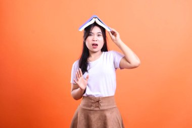 Bir kadın açık bir kitabı şaşırmış bir ifadeyle, beyaz tişört ve kahverengi etek giyen, turuncu arka planda izole edilmiş bir el kaldırmasıyla dengeler.