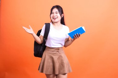 Siyah sırt çantalı genç bir kadın mavi bir kitap tutuyor. Beyaz bir gömlek ve kahverengi bir etek giyiyor ve turuncu arka planda sırıtıyor.