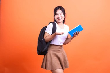 Siyah bir sırt çantası ve mavi bir kitapla turuncu bir arka plana poz veren bir öğrenci..