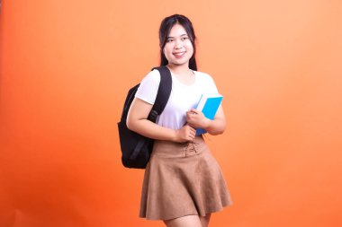 Sırt çantası ve mavi kitabı olan genç bir kadın kameraya gülümsüyor. Beyaz tişört ve kahverengi etek giyiyor. Turuncu arka planda izole edilmiş.