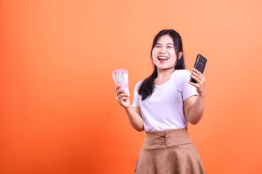 Bir elinde bir yığın para, diğer elinde akıllı bir telefon cihazı olan bir kadın turuncu bir arka plana gülümsüyor. turuncu arkaplanda izole