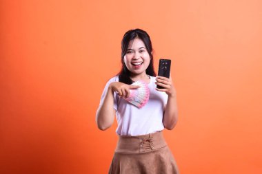 Bir insan elinde bir tomar para ve cep telefonu tutar, gülümser ve önüne bakar. Koyu renk saçlı, beyaz tişörtlü ve kahverengi etekli, turuncu arka planda soyutlanmış..