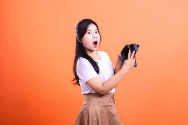 Şaşırmış bir kadının elinde boş siyah bir cüzdan var. Şok olmuş görünüyor. Arka plan katı turuncu. Beyaz tişört ve kahverengi etek giyiyor, turuncu arka planda izole edilmiş..