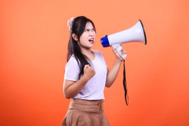 Bir kadın sağ elinde beyaz ve mavi bir megafonla bağırıyor, yumruğunu sol eline dayamış, turuncu bir arka planda soyutlanmış..