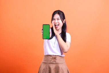 Gülümseyen bir kadın yeşil ekranlı bir telefonu tutarken diğer elini ağzının yanına koyar. Sanki bağırır gibi, turuncu arka planda izole bir şekilde..