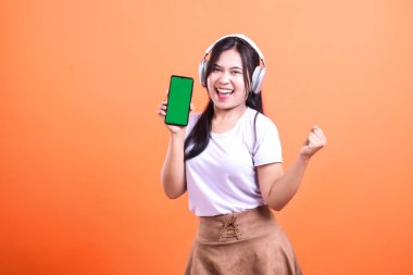 Beyaz tişört ve kahverengi etek giyen bir kadın yeşil ekran akıllı telefonu tutarken gülümsüyor ve turuncu arka planda sağlam bir kulaklık dinliyor..