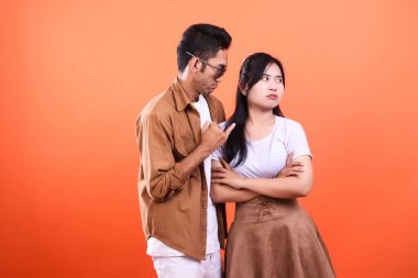 Turuncu arka planda duran genç bir çift. Adam, kollarını kavuşturup yüzünü çeviren kadını işaret ediyor. Anlaşmazlık içindeler..