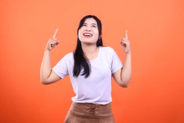 Bir kadın iki eliyle yukarıya bakıyor. Uzun siyah saçları, beyaz bir bluzu ve kahverengi bir eteği var. Gülümsüyor ve turuncu bir arka planda izole edilmiş..