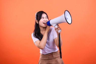 Genç bir kadın elinde beyaz ve mavi bir megafon tutuyor ve ona bağırıyor. O, beyaz bir gömlek ve kahverengi bir etek giyiyor. Turuncu bir arka planda izole edilmiş..
