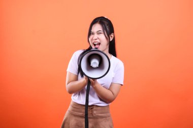 Genç bir kadın iki eliyle megafon tutuyor. Canlı turuncu arka planda izole bir şekilde kameraya bakıyor ve bağırıyor..