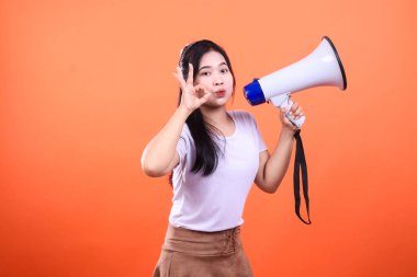 Siyah saçlı bir kadın, tamam işareti yaparken beyaz ve mavi bir megafon tutuyor. Açık mor bir gömlek ve kahverengi bir etek giyiyor. Arka plan turuncu..