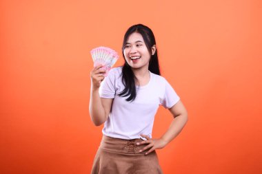 Elinde turuncu arka planda pembe banknotlar tutan gülümseyen bir kadın portre için poz veriyor.