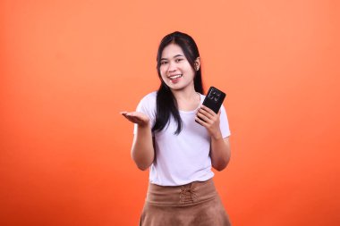 Uzun siyah saçlı genç bir kadın bir elinde akıllı bir telefon tutar ve diğer eli açık, turuncu bir arka planda izole bir şekilde el kol hareketi yapar. O beyaz bir tişört ve kahverengi bir etek giyiyor..