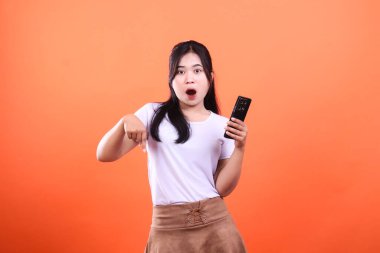 Şaşırmış bir kadın elinde telefon tutarken onu işaret ediyor. Uzun koyu saçlı, beyaz bluzlu ve kahverengi etekli. Arkaplan turuncu bir renktir, model turuncu bir arkaplanda izole edilmiştir.